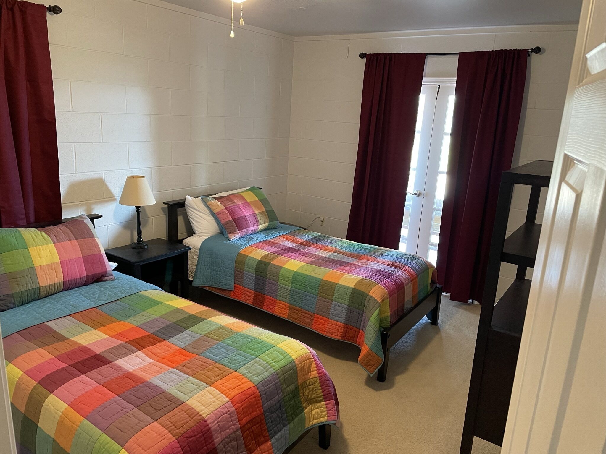 A room with two beds, each with a colorful quilt, a nightstand with a lamp, and a window with curtains.