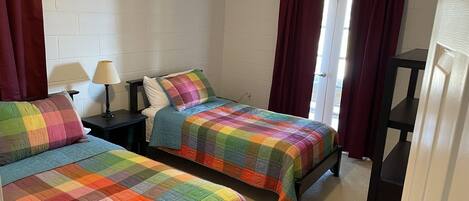 A room with two beds, each with a colorful quilt, a nightstand with a lamp, and a window with curtains.