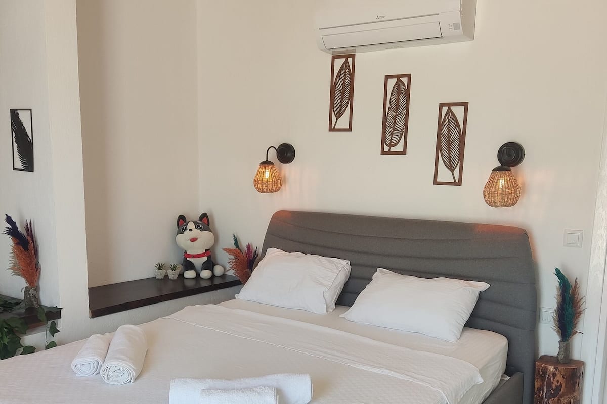 Una cama bien tendida con sábanas blancas y toallas enrolladas, un cabecero tapizado en gris y un aire acondicionado instalado en la pared.