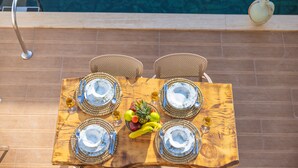 A wooden dining table with four plates, cutlery, and a fruit bowl, set against a poolside backdrop. - Villa Ay (Kas)