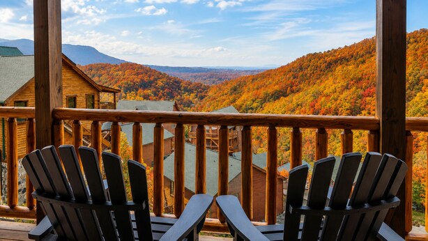 En träveranda med Adirondack-stolar med utsikt över ett bergigt landskap i höstfärger.