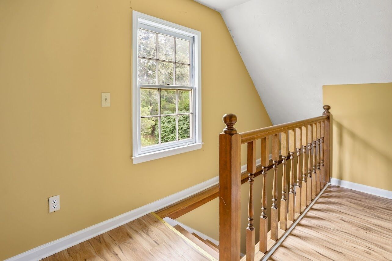 Une pièce avec un escalier en bois, une fenêtre donnant sur la verdure et un mur jaune.