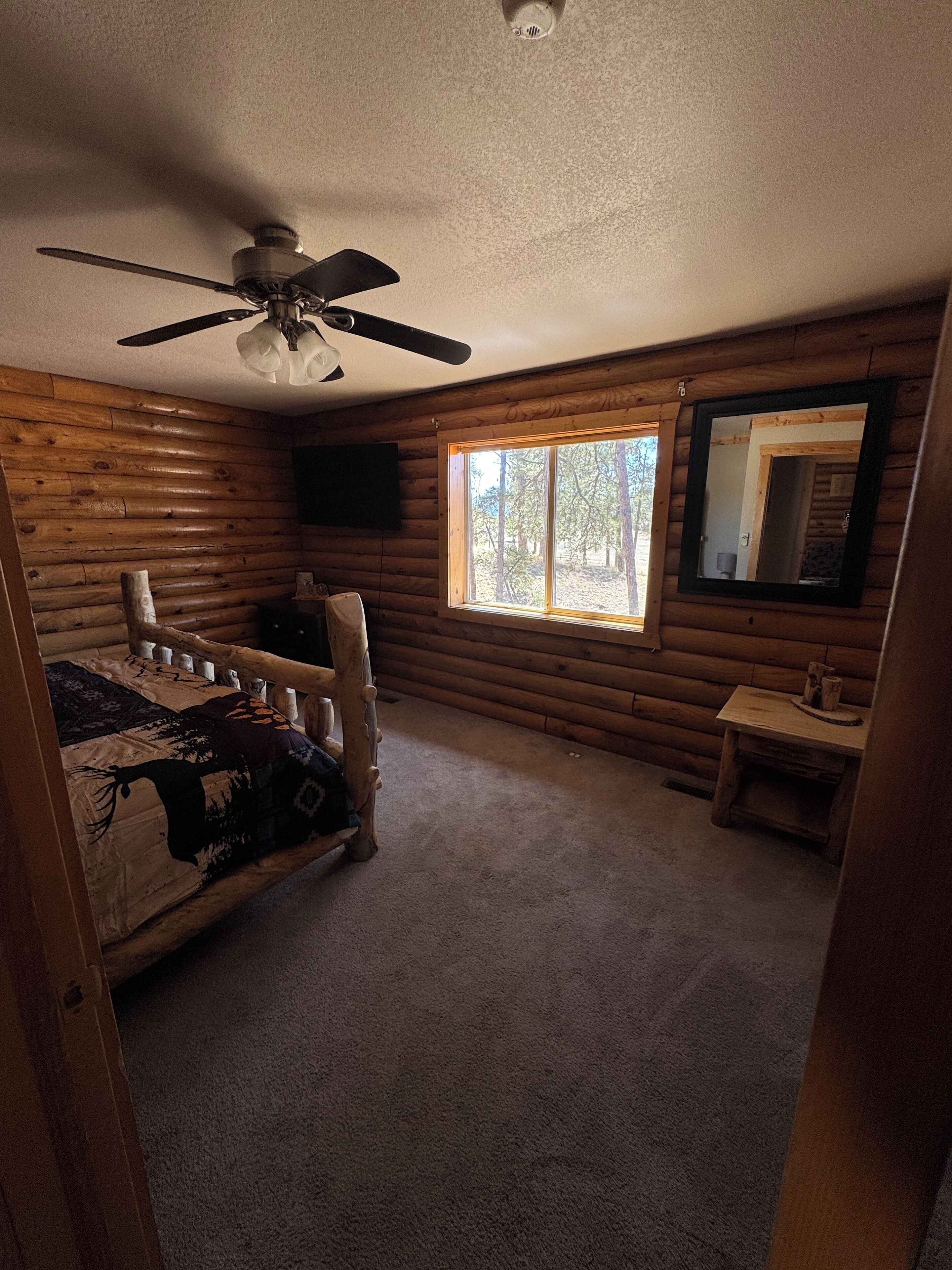 Una habitación tipo cabaña de madera con una cama, un ventilador de techo, una ventana y un espejo.