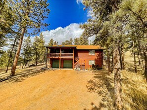 A two-story log cabin with a balcony, surrounded by pine trees.