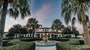 Un jardín bien cuidado con una fuente central, palmeras y un edificio blanco de tejado a la vista.