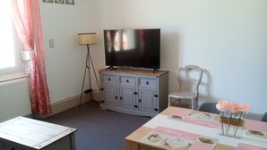 Una habitación con televisión sobre un mueble, una mesa con manteles individuales, una silla y una lámpara.