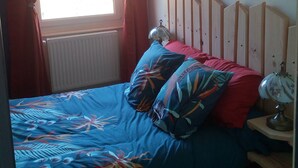 Un dormitorio con cama, cortinas rojas, ventana, radiador, mesita de noche con lámpara y cabecero de madera.