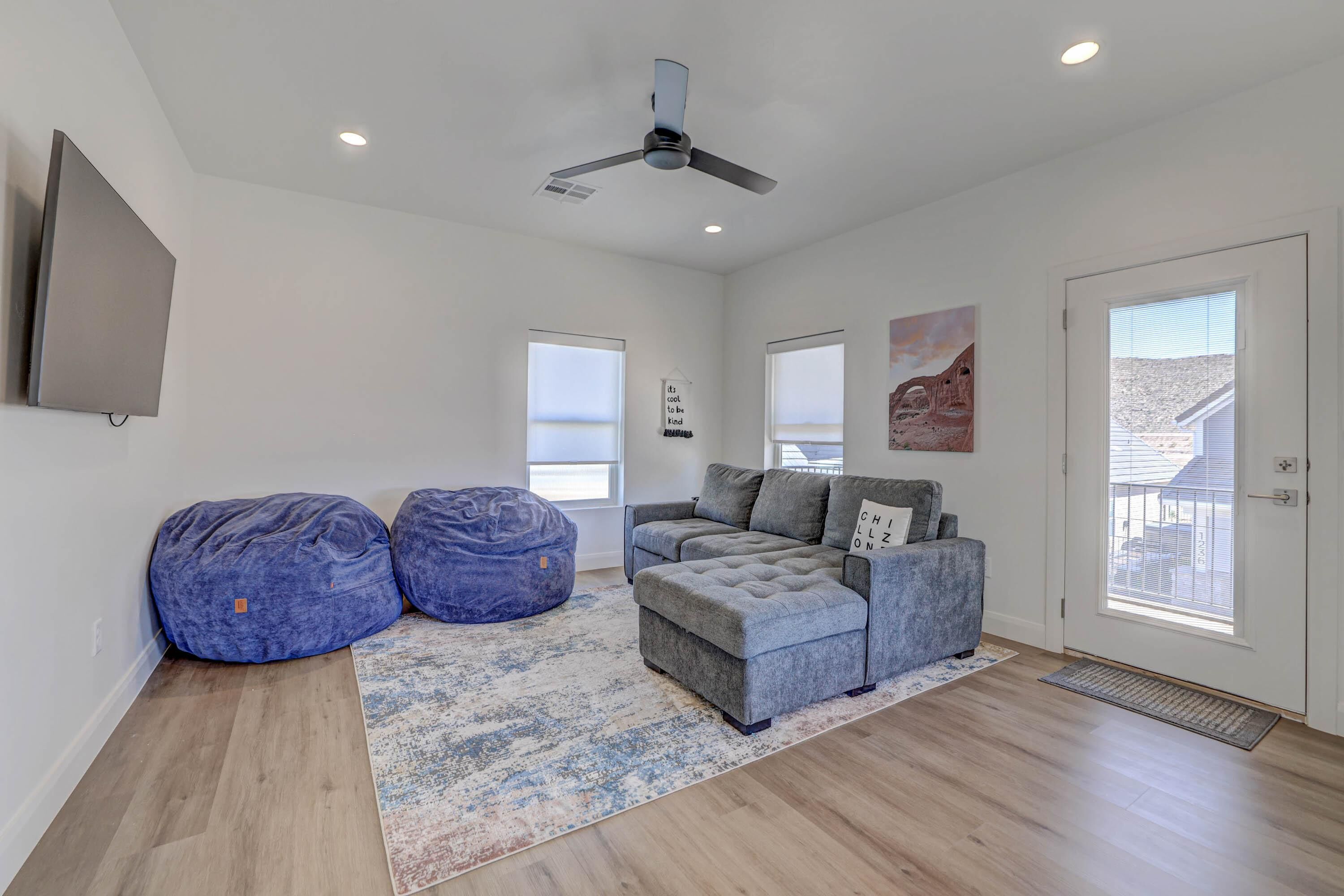 Un salon spacieux avec un canapé d’angle gris, deux fauteuils poire bleus, un ventilateur de plafond et de grandes fenêtres donnant sur l’extérieur.
