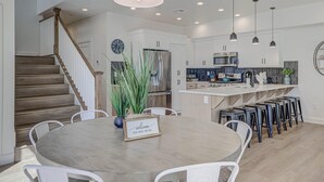 A modern kitchen with white cabinets, a built-in microwave, and a stainless steel refrigerator. There is a wooden staircase leading to another level, and a round dining table with a welcome sign.