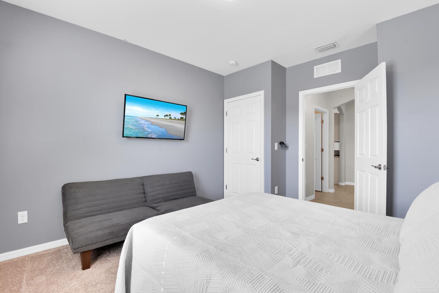 Un dormitorio con un sofá gris, una cama con cubrecama blanco, un televisor empotrado mostrando una escena de playa y una puerta que da a otra habitación.