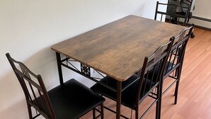 A rectangular wooden dining table with four chairs, each with a black cushioned seat and backrest. The table is positioned against a plain wall, and the floor is covered with wooden planks. - The Village - Private apartment w/ parking 5 min walking (Mamaroneck)
