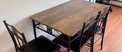 Una mesa rectangular de madera para comer con cuatro sillas, cada una con un asiento y respaldo acolchados en negro. La mesa está colocada contra una pared lisa, y el suelo está cubierto de tablas de madera.