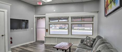 A modern interior with a gray couch, a small wooden table, and a large window offering a view of the street.