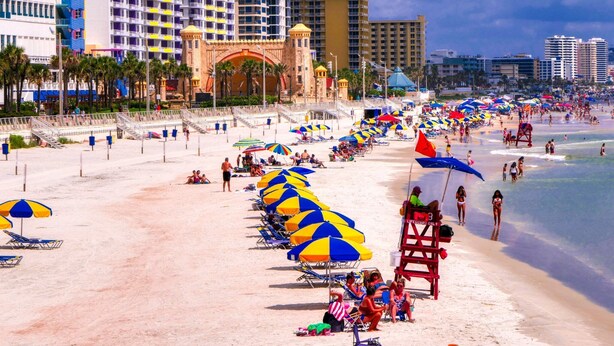 A beach with rows of beach umbrellas and lounge chairs, a lifeguard tower, and a large building with a distinctive archway in the background. - Ocean View Modern Remodeled in Pirates Cove, Full Stove, Balcony Closed. (Daytona Beach Shores)