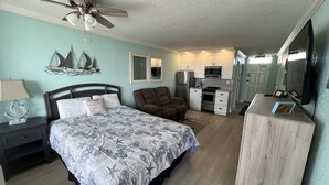 A bedroom with a bed, a chair, a desk, a mirror, a ceiling fan, and a kitchenette. - Ocean View Modern Remodeled in Pirates Cove, Full Stove, Balcony Closed. (Daytona Beach Shores)