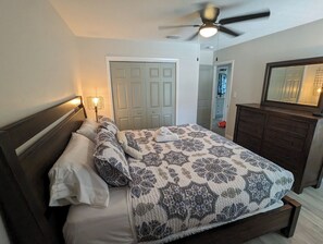 A bedroom with a bed, a ceiling fan, a nightstand with a lamp, a dresser, and a door leading to another room. - Cozy Retreat w/ Heated Pool & Yard Near Downtown (Tampa)