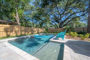 A rectangular swimming pool with a wooden deck surrounding it, a wooden fence, and a large tree in the background.