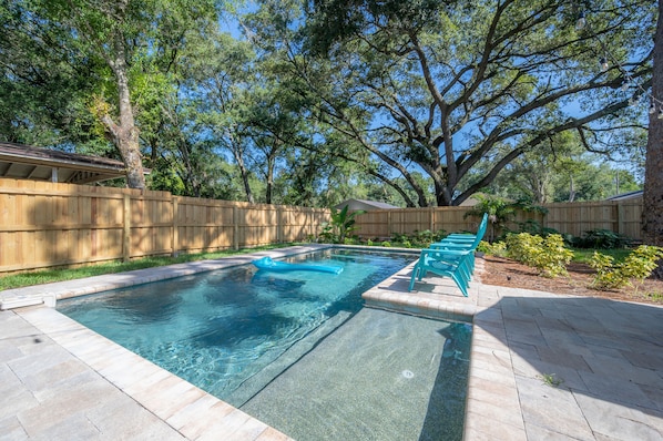 A rectangular swimming pool with a wooden deck surrounding it, a wooden fence, and a large tree in the background. - Cozy Retreat w/ Heated Pool & Yard Near Downtown (Tampa)
