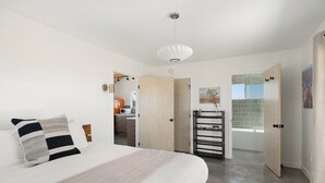 A bedroom with a bed, a bedside table, a hanging light fixture, and a view of a bathroom with a bathtub. - Green Jackrabbit - Hot Tub + Endless Desert Views! (Joshua Tree)