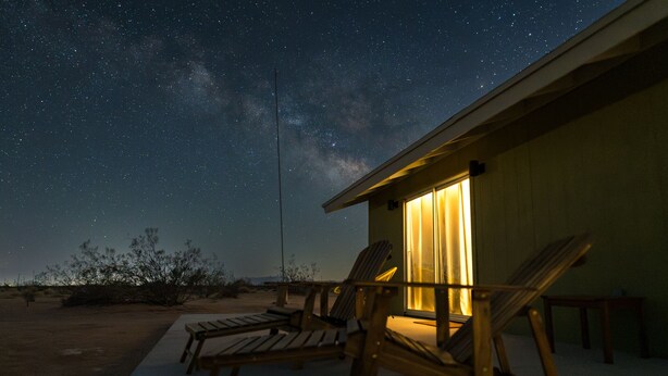 A starry night sky with a view of the Milky Way, a wooden deck with two lounge chairs, and a building with a lit interior.
