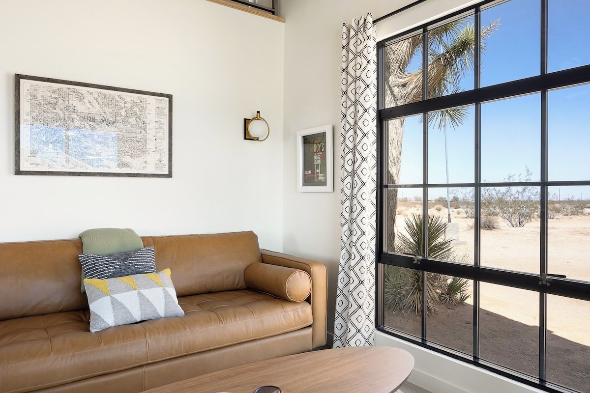 A room with a large window, a brown leather sofa, a framed map, and a framed picture on the wall.