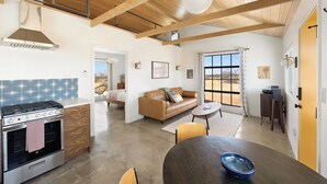 A modern kitchen with a stove, oven, and wooden ceiling beams. There is a dining area with a table and chairs, and a living space with a sofa and a window. - Green Jackrabbit - Hot Tub + Endless Desert Views! (Joshua Tree)