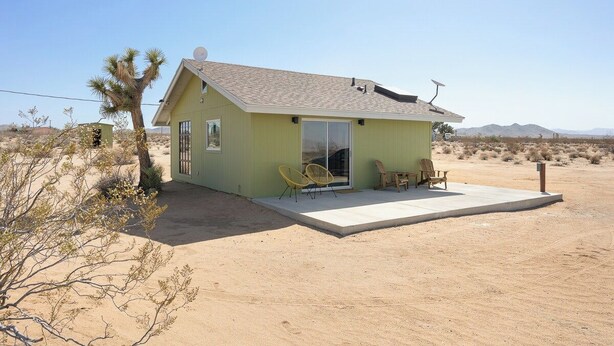 A small, single-story house with a flat roof, a patio with chairs, and a desert landscape in the background. - Green Jackrabbit - Hot Tub + Endless Desert Views! (Joshua Tree)