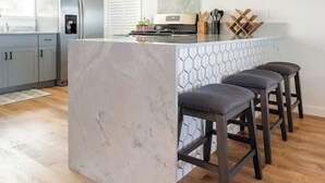 A modern kitchen with a marble island, bar stools, stainless steel appliances, pendant lighting, and a large window. - Hale 'Io • Remodeled 2/2 Luxury • AC • Ocean View (Kailua-Kona)