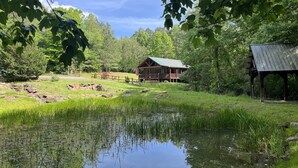A cabin with a green roof, surrounded by a pond, trees, and a wooden fence.