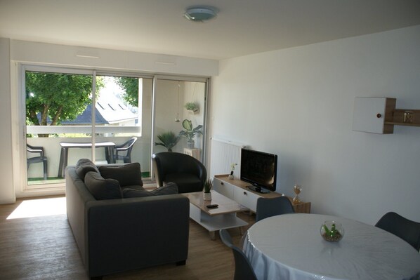 Ein modernes Wohnzimmer mit Sofa, Fernseher, Esstisch und einem Balkon mit Gartenmöbeln.