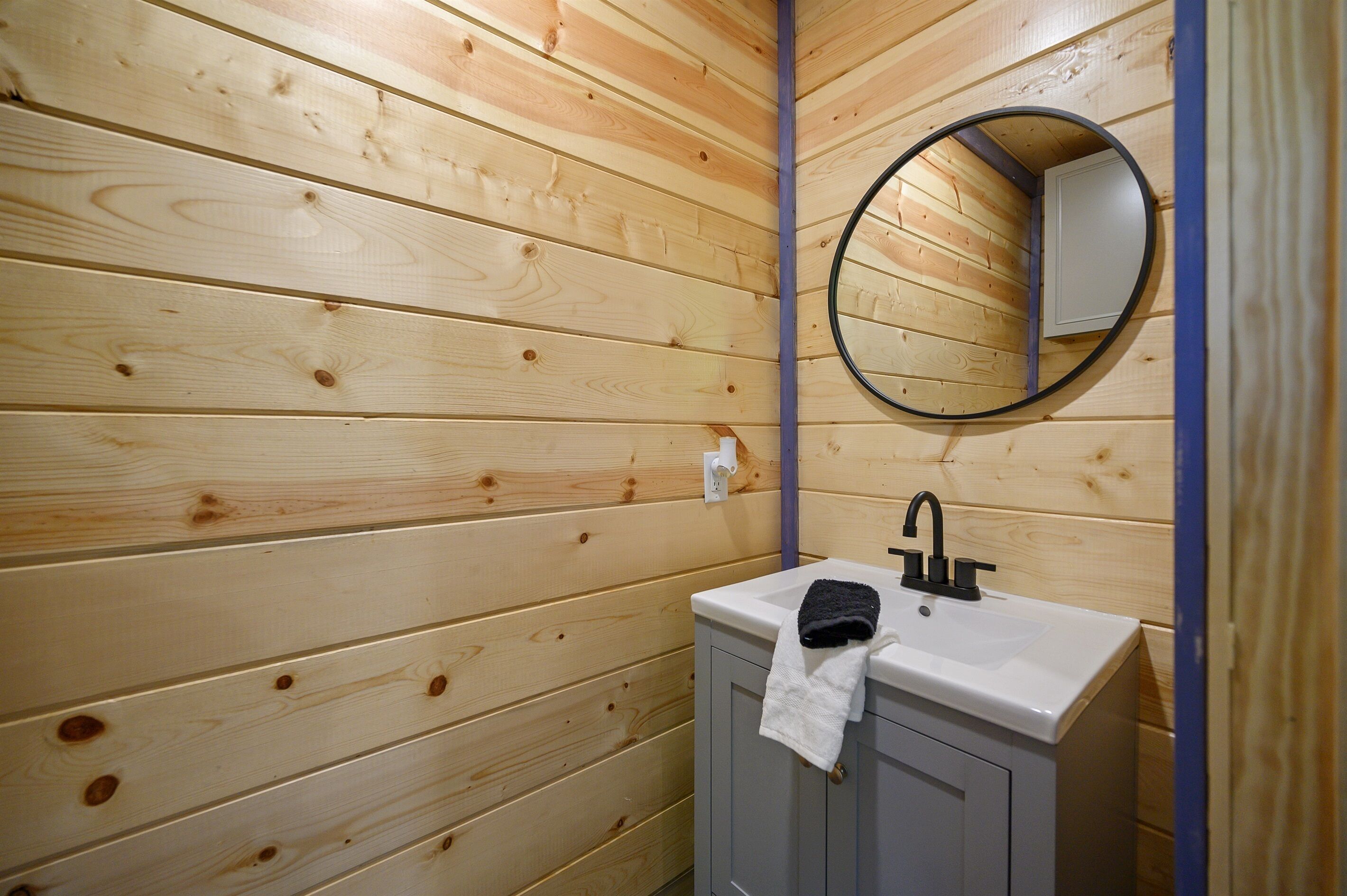 Une salle de bain aux murs lambrissés, avec un lavabo blanc, un robinet noir et un miroir rond.