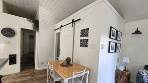 Un comedor con una mesa de madera y sillas blancas, un reloj negro y cuadros en la pared.