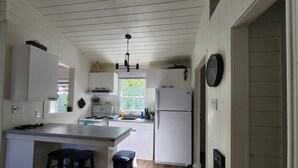 Una cocina con armarios blancos, un lavamanos doble, una estufa y un refrigerador. Hay dos taburetes y un cubo de basura.