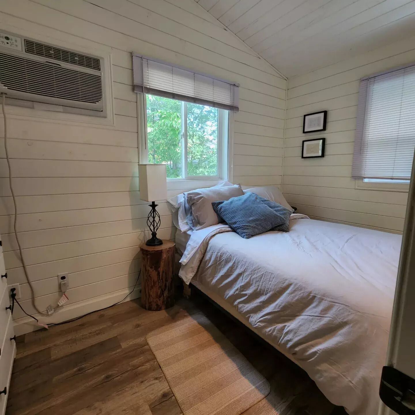 A bedroom with a bed, a window with blinds, a wall-mounted air conditioner, and a small wooden stool with a lamp.