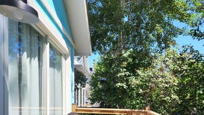 Una terraza de madera con dos sillas y una mesa, barandilla y un edificio con detalles azules y ventanas amplias.