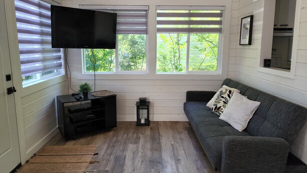 Un salón con un sofá gris, un televisor de pantalla plana montado en la pared, un mueble de entretenimiento negro pequeño y una ventana con persianas.