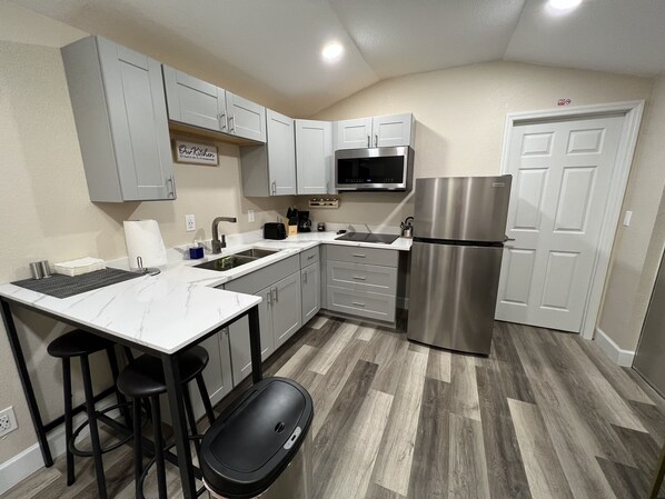 Una cocina moderna con armarios grises, un refrigerador de acero inoxidable, un microondas y un lavabo. Hay taburetes de bar y una encimera con dispensador de toallas de papel.
