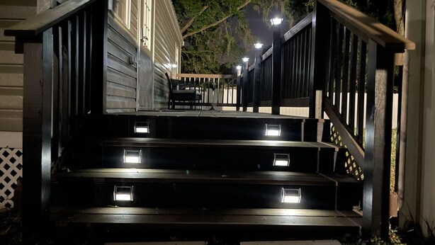 Degraus de madeira ao ar livre levam a uma casa com varanda, iluminada por luzes.