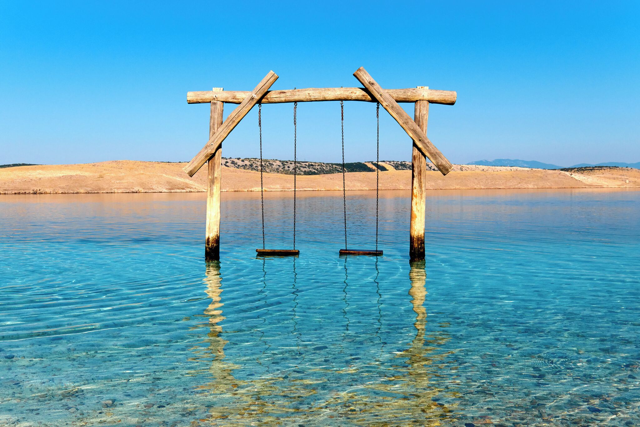 A wooden swing set over clear water with a sandy beach in the background.