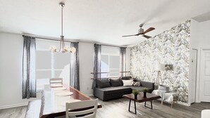 Ein geräumiges Wohnzimmer mit Essbereich, grauem Sofa, Deckenventilator und einer Wand mit floralem Wandpapier.