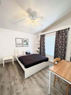 Un dormitorio con cama, mesita de noche, una silla, mesa, ventilador de techo y ventana con cortinas.
