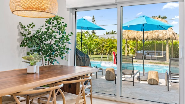 A modern room with a wooden dining table, wicker chairs, a large window with a view of a pool and outdoor furniture. - Casa Del Sol - Bike to Beach, Saltwater Pool & Private Backyard (Palm Beach Gardens)