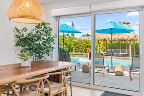 Une pièce moderne avec une table à manger en bois, des chaises en osier, une grande fenêtre donnant sur une piscine et du mobilier d’extérieur.