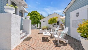 A patio with white furniture and planters, a brick floor, and a white building with a window and a door.