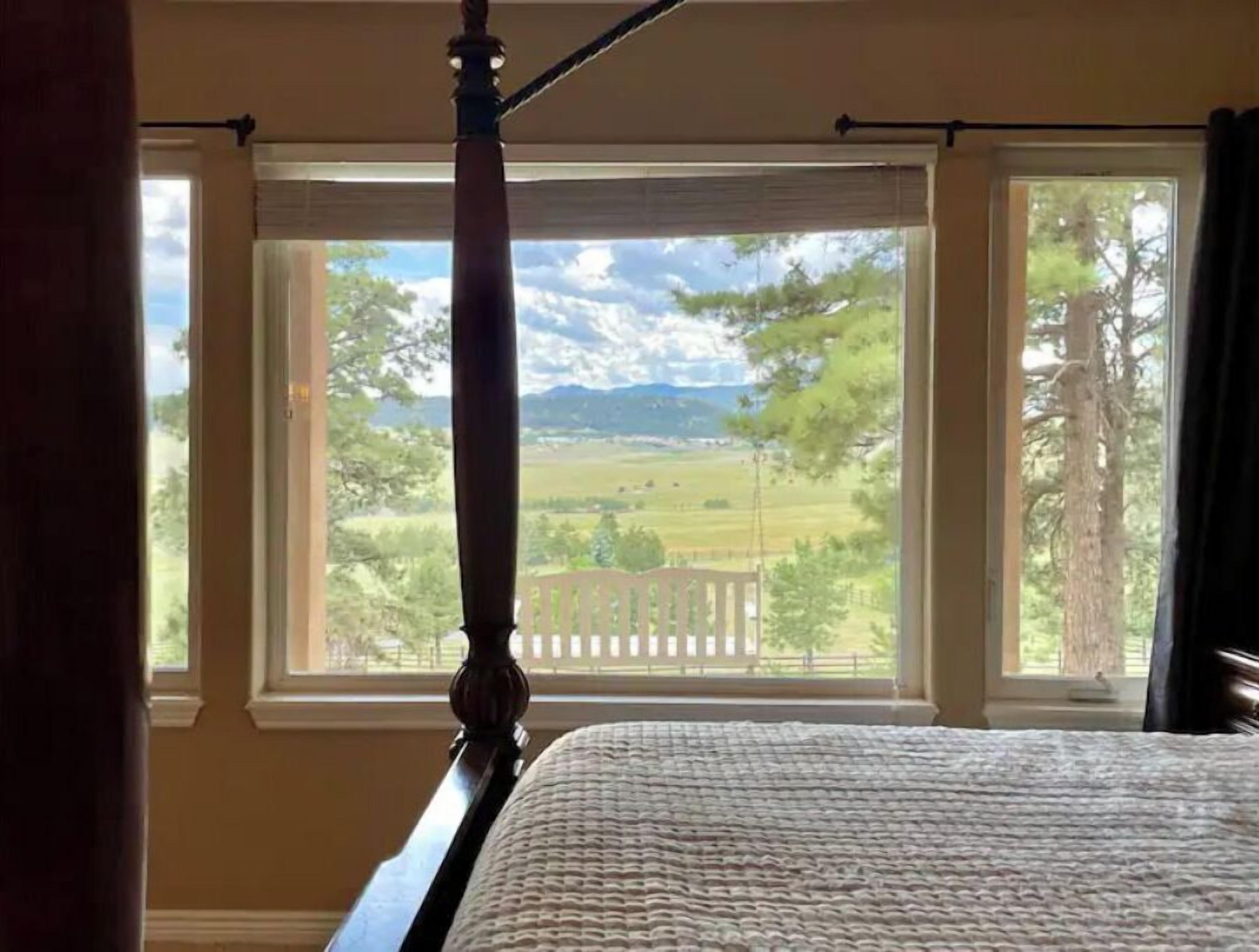 Un dormitorio con cama con dosel, ventanas amplias con vista a un paisaje rural y un cabecero de madera.