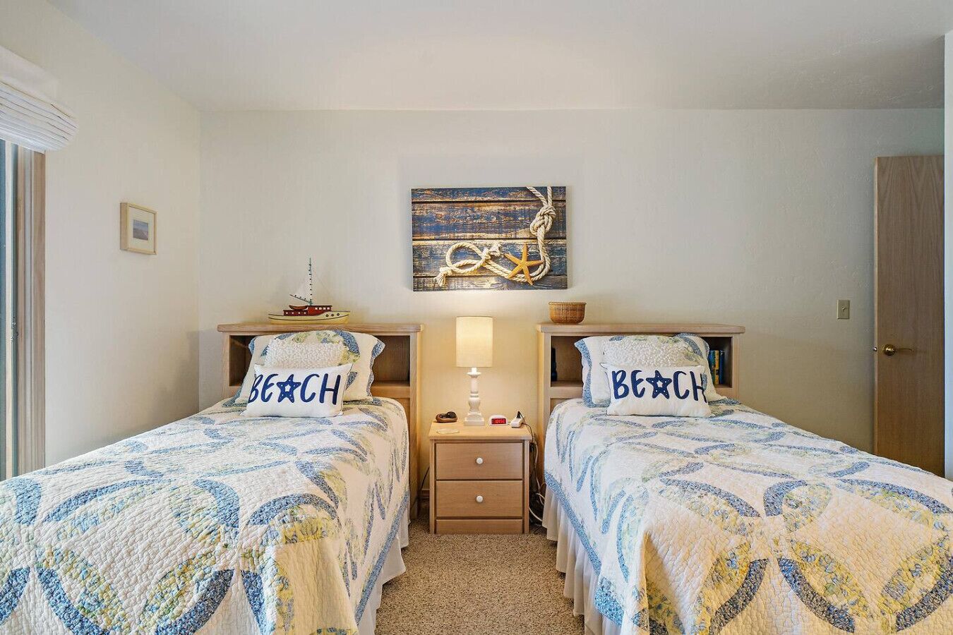 Een kamer met twee bedden, elk voorzien van een kussen met de tekst 'BEACH', een klein nachtkastje met een lamp, en een decoratie aan de muur boven de bedden.