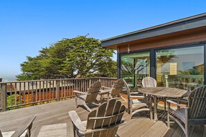 Una terraza con sillas y una mesa de madera, con vista a un árbol y un edificio con ventanas grandes.