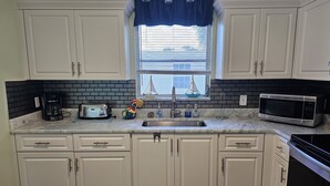 Una cocina con armarios blancos, salpicadero blanco y negro, un ventanal con decoración de velero y un microondas de acero inoxidable.