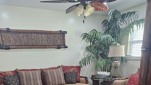 Una sala de estar con una mesa de centro de vidrio, un sofá, un tapete estampado, un ventilador de techo y una planta grande.