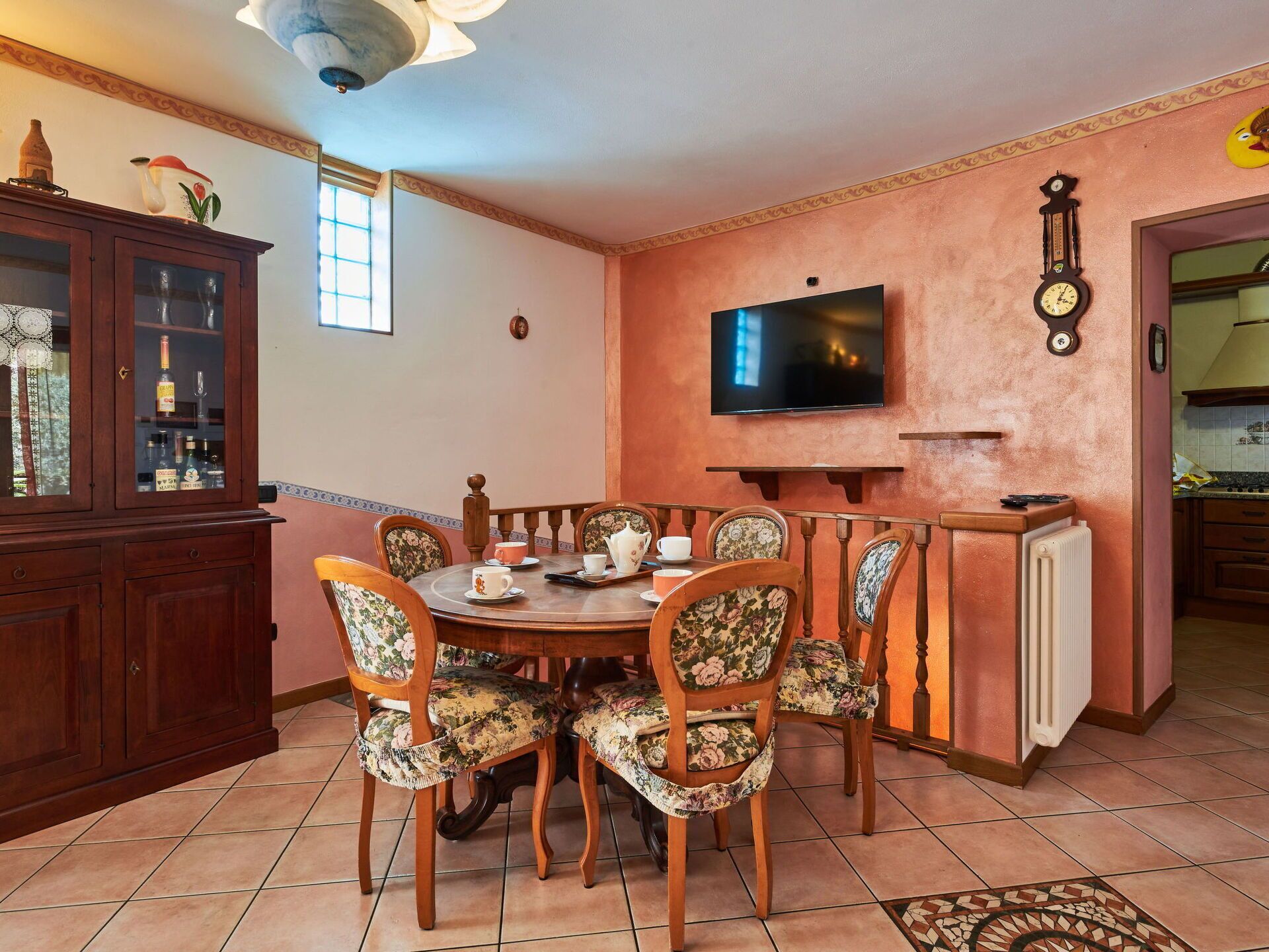 Un comedor con una mesa redonda, sillas con motivos florales, un televisor montado en la pared y un mueble con puertas de cristal.
